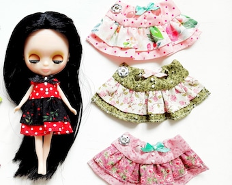 May include: Six colorful doll skirts with floral and polka dot patterns. Each skirt has three layers of fabric and a bow. The skirts are arranged around a doll with black hair and a red and black polka dot dress.
