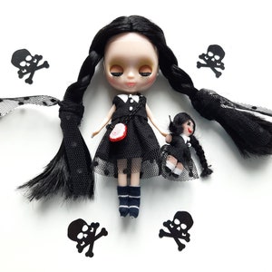 May include: A black doll with long black braids wearing a black dress with white polka dots and black lace. The doll is holding a smaller black doll with black hair and a black dress. The dolls are surrounded by black skull and crossbones cutouts.