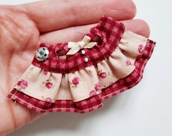 May include: A miniature dress with a red and white gingham pattern and a floral print. The dress has a ruffled skirt and a bow at the neckline.