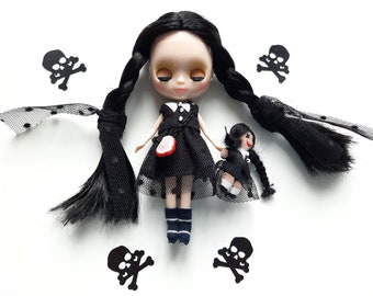 May include: A black doll with long black braids wearing a black dress with white polka dots and black lace. The doll is holding a smaller black doll with black hair and a black dress. The dolls are surrounded by black skull and crossbones cutouts.