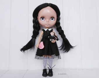 May include: A black-haired doll wearing a black dress with white polka dots and white and black striped tights. The doll is holding a smaller doll in a black dress. The doll is standing on a white wooden surface.