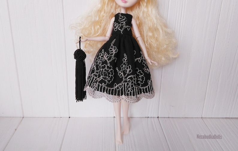 May include: A black tulle dress with silver embroidery and a scalloped lace trim. The dress is being worn by a doll with blonde hair and is holding a black tassel.