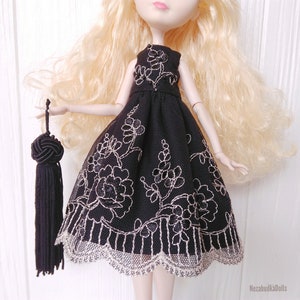 May include: A black tulle dress with silver floral embroidery and a lace trim. The dress is being worn by a doll with blonde hair and a black tassel.