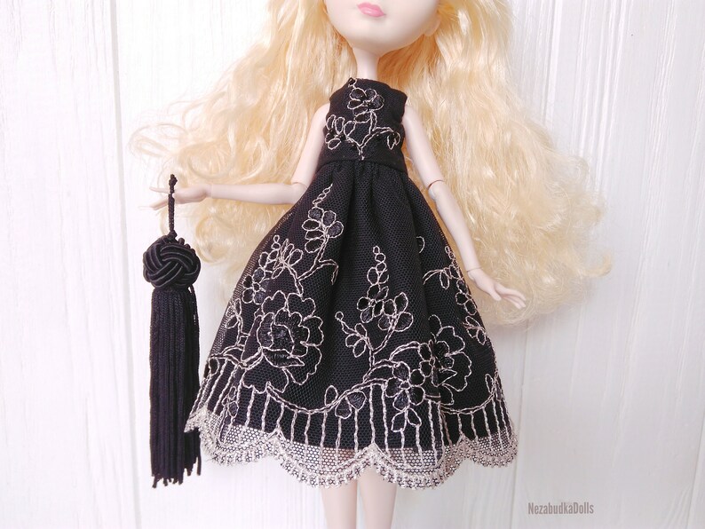 May include: A black tulle dress with silver floral embroidery and a lace trim. The dress is being worn by a doll with blonde hair and a black tassel.