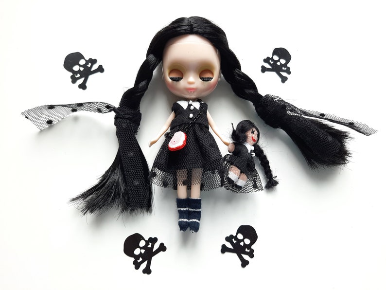 May include: A black doll with long black braids wearing a black dress with white polka dots and black lace. The doll is holding a smaller black doll with black hair and a black dress. The dolls are surrounded by black skull and crossbones cutouts.