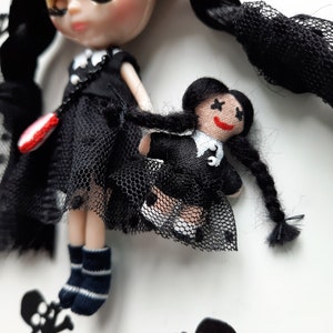 May include: Two black-haired dolls wearing black dresses with white collars and black mesh skirts. The larger doll has a red and white heart-shaped accessory. The smaller doll has a black and white cross on her dress.