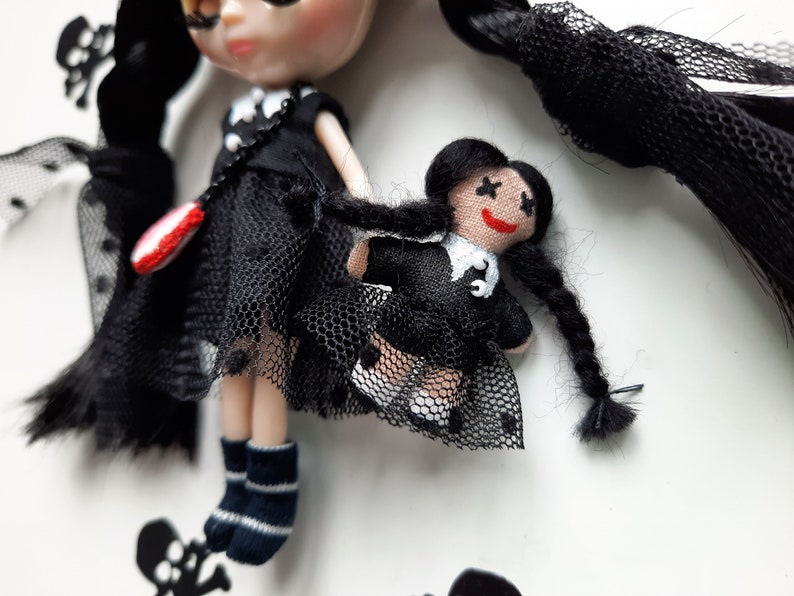 May include: Two black-haired dolls wearing black dresses with white collars and black mesh skirts. The larger doll has a red and white heart-shaped accessory. The smaller doll has a black and white cross on her dress.