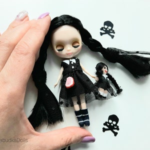 May include: Two black-haired dolls wearing black dresses and white socks. The larger doll has a red heart-shaped accessory on her dress. Both dolls have black braids. The dolls are surrounded by black paper cutouts of skulls and crossbones.