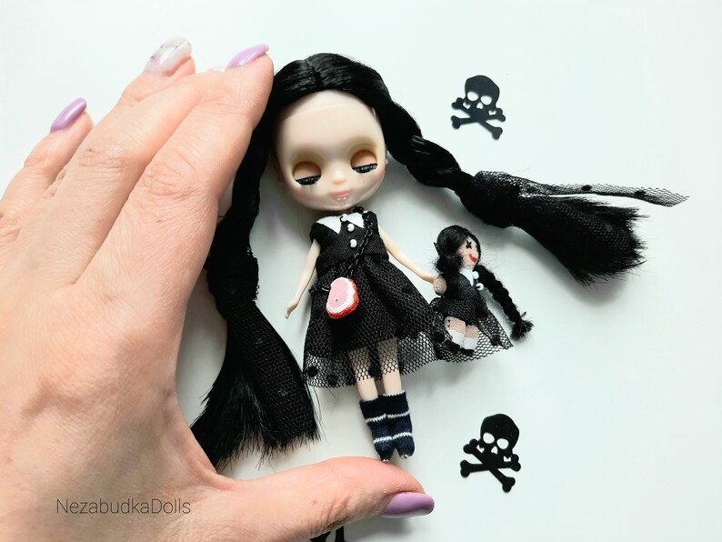 May include: Two black-haired dolls wearing black dresses and white socks. The larger doll has a red heart-shaped accessory on her dress. Both dolls have black braids. The dolls are surrounded by black paper cutouts of skulls and crossbones.