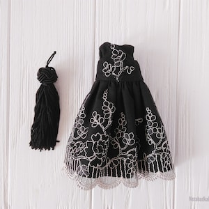 May include: A black dress with a white floral embroidery design and a lace trim. The dress is on a white wooden surface.