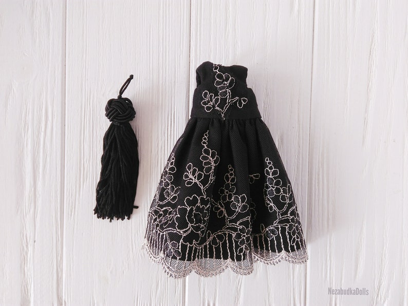 May include: A black dress with a white floral embroidery design and a lace trim. The dress is on a white wooden surface.
