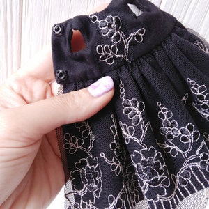 May include: Black tulle dress with silver embroidery of floral designs. The dress has a scalloped lace trim and two black buttons on the shoulder straps.
