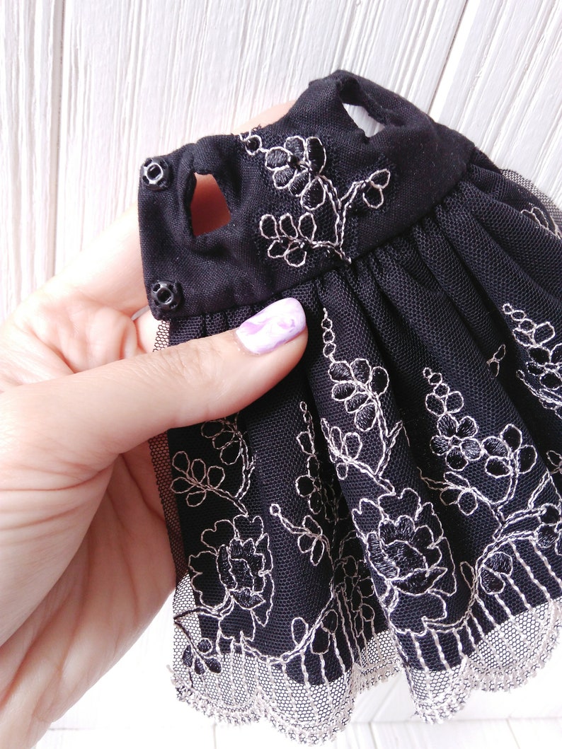 May include: Black tulle dress with silver embroidery of floral designs. The dress has a scalloped lace trim and two black buttons on the shoulder straps.