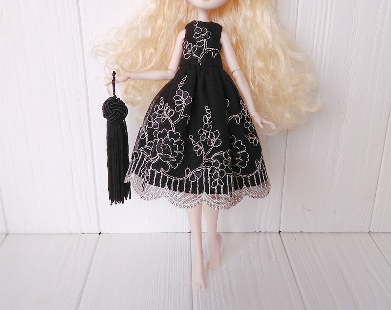 May include: A black dress with silver floral embroidery and a lace trim. The dress is being worn by a doll with blonde hair.