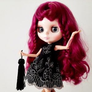 May include: A doll with long, curly, burgundy hair wearing a black dress with white embroidery and holding a black tassel.