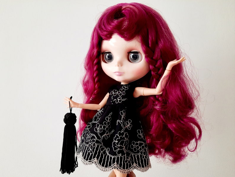 May include: A doll with long, curly, burgundy hair wearing a black dress with white embroidery and holding a black tassel.