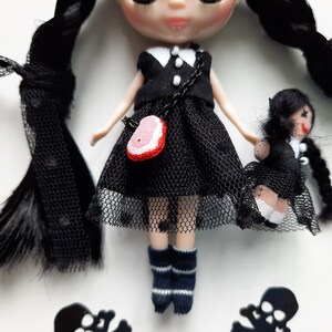 May include: A black and white doll with long black hair wearing a black dress with a white collar and a red and white purse. The doll is holding a smaller black and white doll with long black hair wearing a black dress with a white collar.