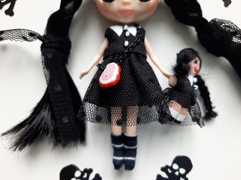 May include: A black and white doll with long black hair wearing a black dress with a white collar and a red and white purse. The doll is holding a smaller black and white doll with long black hair wearing a black dress with a white collar.