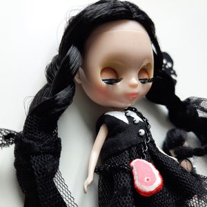 May include: A black haired doll wearing a black dress with a white collar and a necklace with a red meat charm. The doll has long black hair styled in braids and is lying on a white surface.