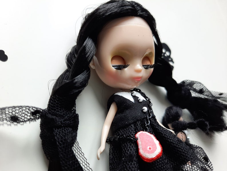 May include: A black haired doll wearing a black dress with a white collar and a necklace with a red meat charm. The doll has long black hair styled in braids and is lying on a white surface.