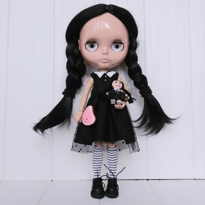 May include: A black-haired doll with large eyes wearing a black dress with white polka dots and white and black striped stockings. The doll is holding a smaller doll in a black dress. The doll is standing on a white wooden surface.