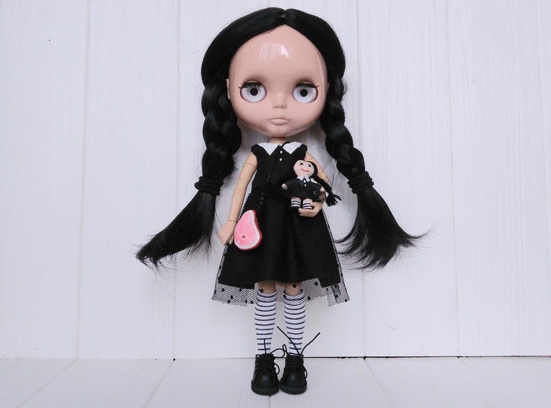 May include: A black-haired doll with large eyes wearing a black dress with white polka dots and white and black striped stockings. The doll is holding a smaller doll in a black dress. The doll is standing on a white wooden surface.