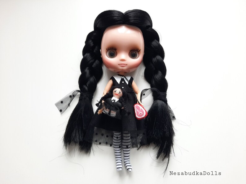 May include: A black haired doll with a black and white dress and black and white striped stockings. The doll is holding a smaller doll in a similar outfit. The doll is wearing a necklace with a small red meat charm.