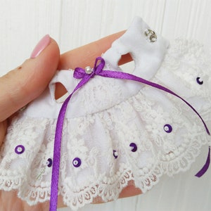 May include: White lace doll dress with purple ribbon and silver embellishments. The dress has a ruffled skirt and a white bodice.