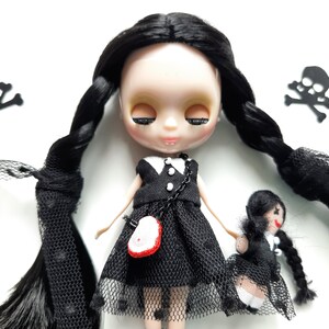 May include: A black doll with long black braids wearing a black dress with white trim and a black mesh overlay. The doll has a white face with closed eyes and a pink mouth. The doll is holding a smaller black doll with a black dress and a white face. There are two black skull and crossbones decorations in the background.