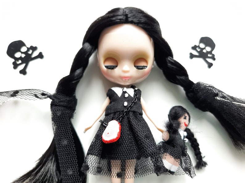 May include: A black doll with long black braids wearing a black dress with white trim and a black mesh overlay. The doll has a white face with closed eyes and a pink mouth. The doll is holding a smaller black doll with a black dress and a white face. There are two black skull and crossbones decorations in the background.