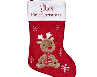 Op de afbeelding: Rode kerstsok met een witte manchet, met de tekst "Ellie's First Christmas". De sok heeft een rendierapplicatie met een rode sjaal en witte sneeuwvlokken. Er is een lus bevestigd om op te hangen.