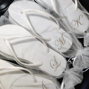 May include: White flip-flops with gold monogram initials, packaged in clear bags tied with white ribbons. The sandals have a textured footbed and a simple thong strap design. A, K, L, and M are visible.