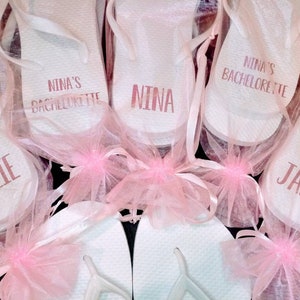 May include: White flip-flops with custom text, including "Nina's Bachelorette", "Nina", "Catlie", and "Jamie". Each pair is wrapped in a sheer pink fabric and tied with a pink ribbon, perfect for a bachelorette party.