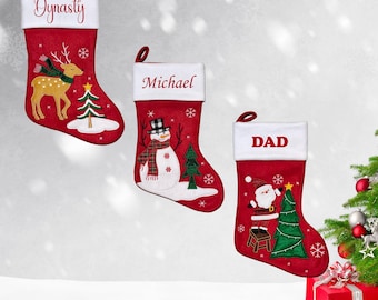 May include: Three red Christmas stockings with white cuffs and hanging loops. Each stocking has a different design: a reindeer, a snowman, and Santa Claus. The stockings are personalized with the names "Dynasty", "Michael", and "DAD".