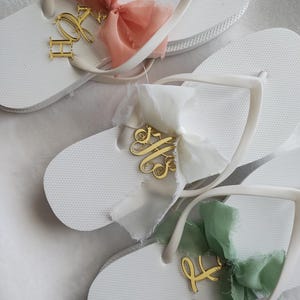 May include: White flip-flops with gold initial charms and colorful ribbons. The sandals feature a textured sole and white straps. The ribbons are in shades of peach, white, and green. The gold charms display elegant cursive initials.