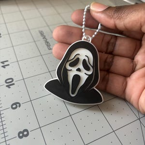 May include: A white and black plastic keychain featuring the iconic Ghostface mask design with a hooded cloak. The keychain is attached to a silver-colored metal ball chain, providing a secure way to attach it to keys, bags, or other items.