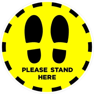 May include: A yellow circle with a black border and two black footprints. The text "PLEASE STAND HERE" is written in black letters.