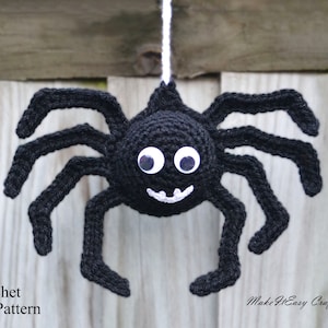 Peut inclure: Une araignée en crochet noir avec des yeux globuleux blancs et une bouche souriante. L'araignée est suspendue à une ficelle blanche sur un fond en bois. Modèle de crochet.