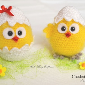 May include: Two crocheted yellow chicks, one in an egg shell and one standing on its own. Both chicks have white eyes, red beaks, and white crocheted hats. The chick in the egg shell has a red bow on its head. The chicks are sitting on a bed of green grass.