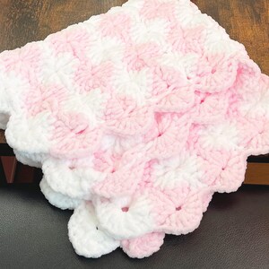 May include: A pink and white crocheted baby blanket with a scalloped edge. The blanket is made with a soft, fluffy yarn.