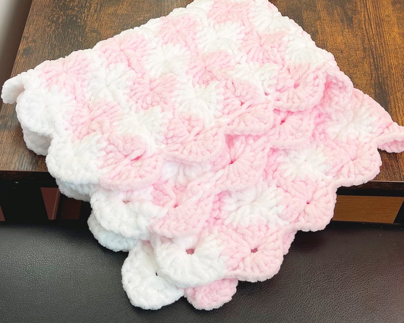 May include: A pink and white crocheted baby blanket with a scalloped edge. The blanket is made with a soft, fluffy yarn.
