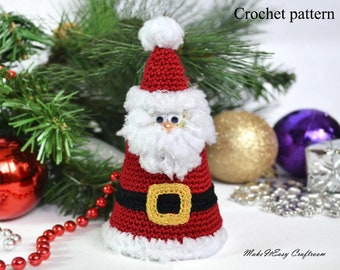 Puede incluir: Adorno de crochet de Santa Claus en rojo y blanco con un cinturón negro y una hebilla dorada. El adorno está sobre una superficie blanca con ramas de pino verdes y adornos rojos y plateados en el fondo.
