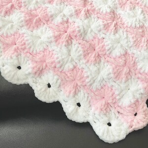 May include: A pink and white crocheted baby blanket with a scalloped edge. The blanket is made with a soft, fluffy yarn and has a textured pattern.