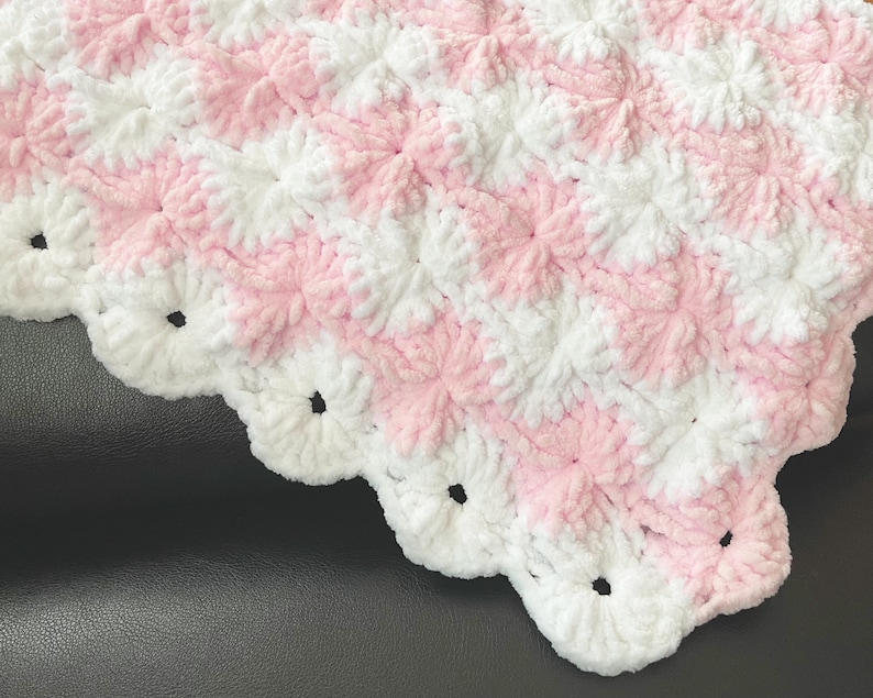 May include: A pink and white crocheted baby blanket with a scalloped edge. The blanket is made with a soft, fluffy yarn and has a textured pattern.