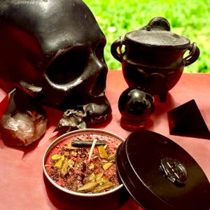 May include: A black skull, a small black cauldron, a black crystal ball, a black pyramid, and a red candle with herbs and crystals inside a black tin.