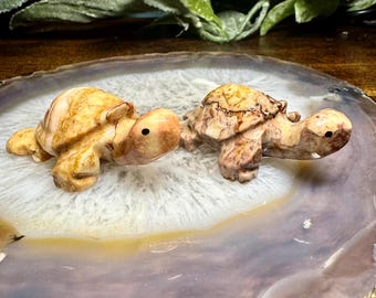 May include: Two small, carved stone turtles in shades of tan and brown are displayed on a polished, agate stone. The turtles have detailed shells and small, dark eyes. The agate stone has a white, tan, and gray pattern.
