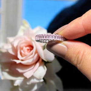 May include: A silver ring with a continuous band of pink rectangular gemstones. The ring is held in front of a soft pink rose and a blue background. The gemstones are evenly sized and set closely together.