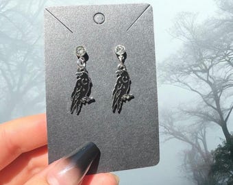 May include: Silver-toned raven earrings with a sparkling gem at the top. Each earring features a raven perched on a branch. The earrings are displayed on a gray card against a misty background.