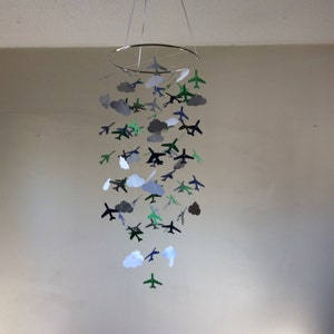 May include: A mobile made of paper airplanes and clouds in shades of gray, green, and blue. The airplanes are suspended from a silver ring.
