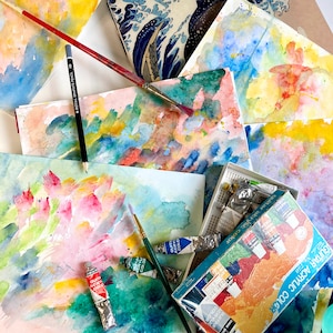 May include: A close-up of a set of watercolour paints with a variety of colours, including blue, green, red, and yellow. The paints are arranged in a box with the words "Guitar Acrylic Color" printed on the front. The box is sitting on a table with several sheets of watercolour paper.
